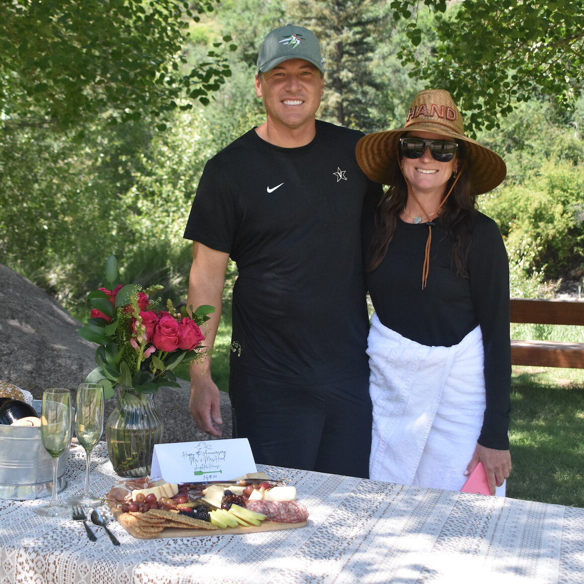 A couple are standing behind a table with roses, champagne, and a charcuterie board with a note wishing them a Happy Anniversary