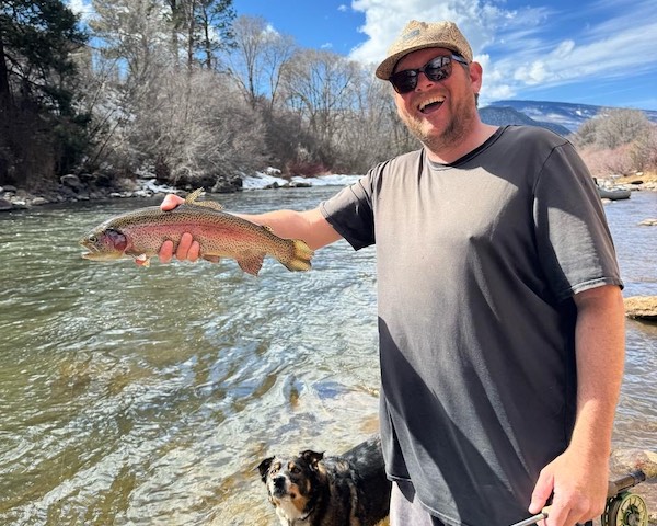Man holding a Rainbow trout by the Roaring Fork River with a dog watching from below