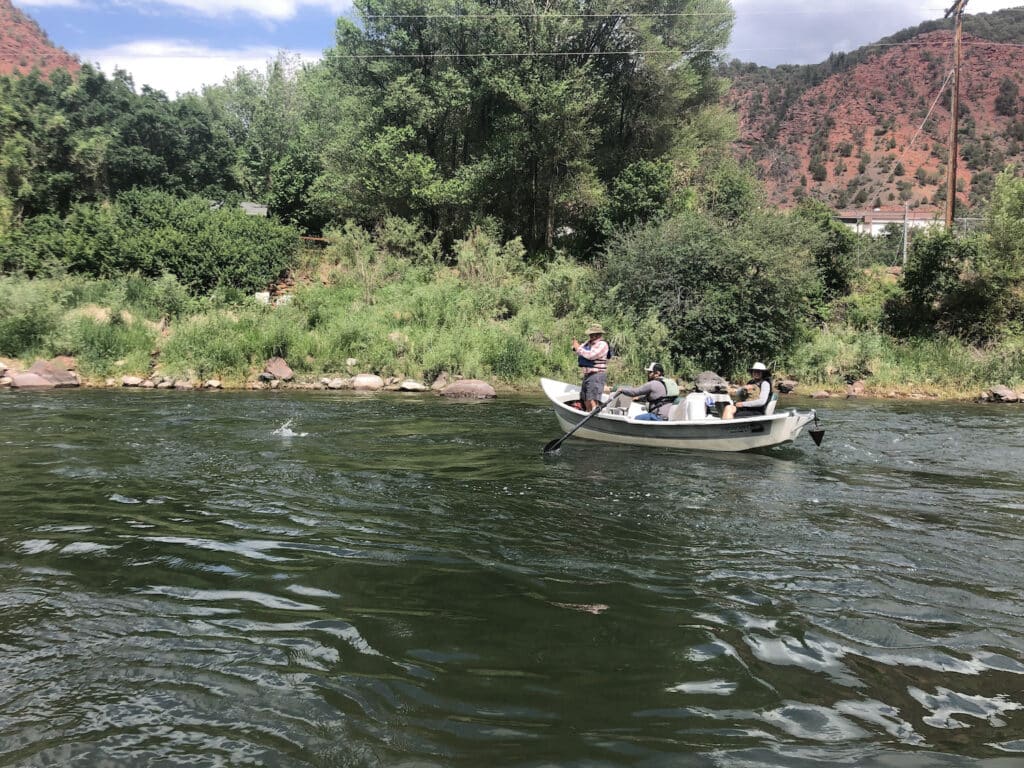 Three people on a drift boat, fly-fishing. Person at the front of the boat has a fish on the line.
